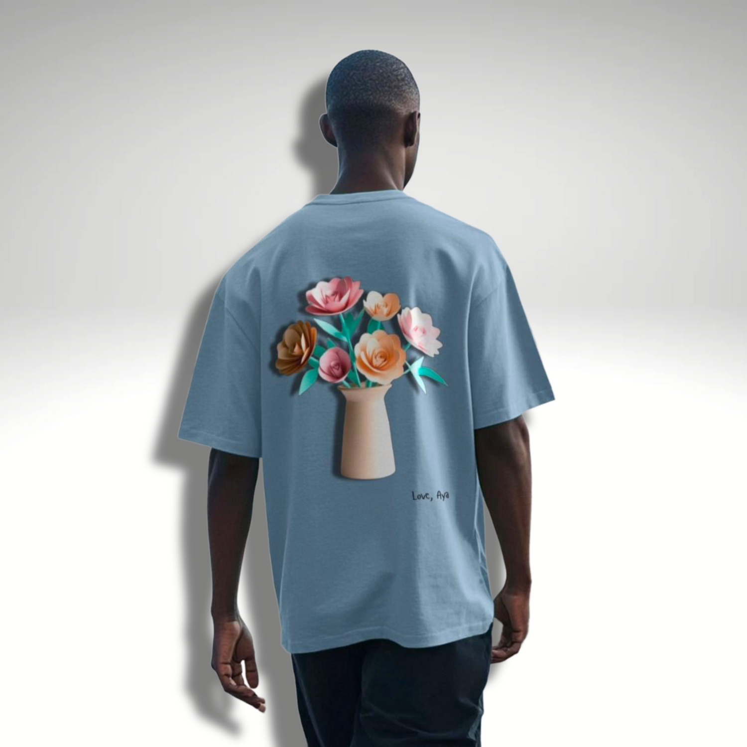 Man facing away from camera modelling an oversized T-Shirt in Stone Blue which has paper flowers in a vase printed on the back. Love, Aya Branded.