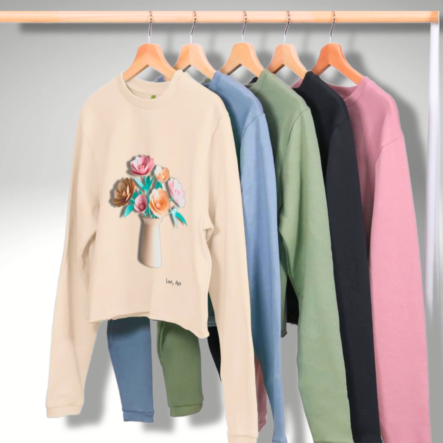 Organic Cropped Jumpers hanging on a rail in various colours. The focal point is the Oat coloured jumper with Paper FLowers in a vase printed on the front. Love, Aya branded.