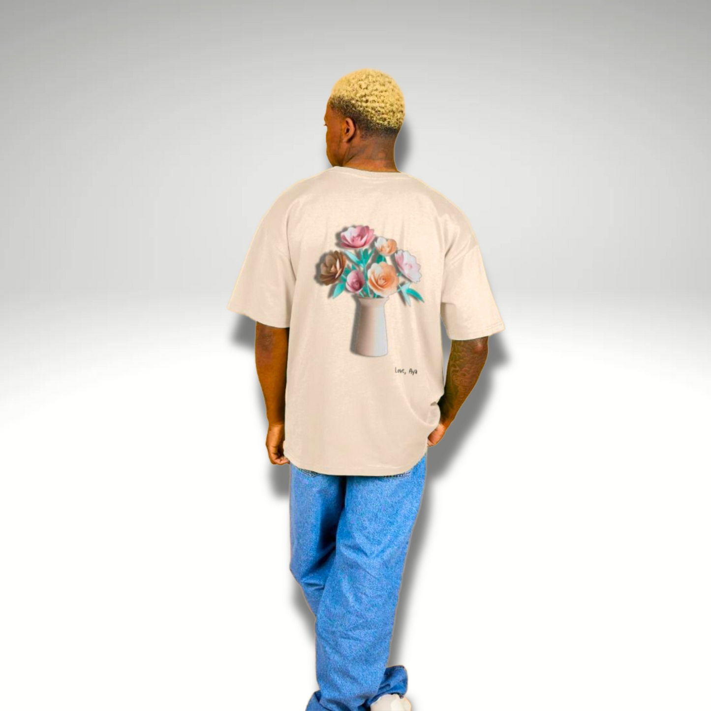 Men’s Oversized T-Shirt in Oat Beige showing the  back of the product which features a large image of paper origami flowers in a vase. It is “Love, Aya” branded and is modelled with blue baggy denim jeans.