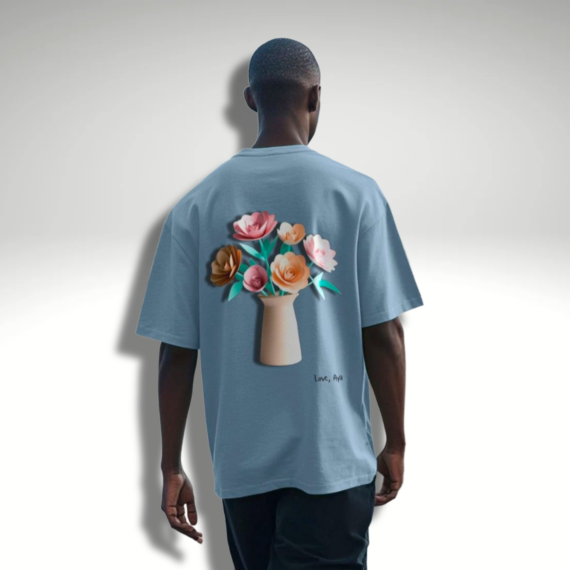 Men’s Oversized T-Shirt in Stone Blue showing the back of the product which features a large image of paper origami flowers in a vase. It is “Love, Aya” branded and is modelled with black baggy denim jeans.